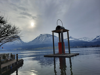 See mit Steg, schwimmender roter Laterne und schneebedeckten Bergen unter hellem, bewölktem Himmel.