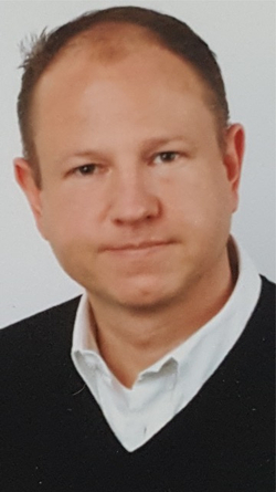 Kopfaufnahme eines Mannes mit kurzen dunkelblonden Haaren und hellbraunen Augen, im weißen Hemd und dunklen V-Ausschnitt-Pullover.