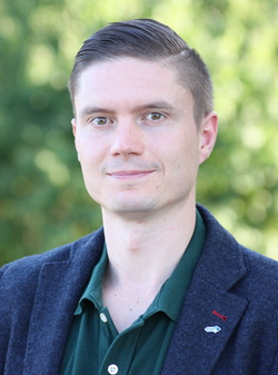 Porträt eines lächelnden jungen Mannes mit kurzen Haaren, dunkelblauem Sakko und grünem Hemd, vor grünem Hintergrund.