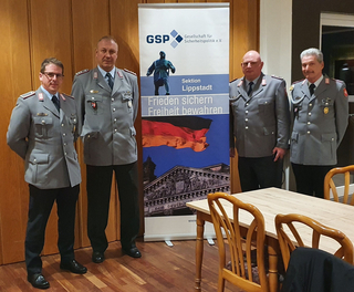 Vier Männer in Bundeswehr-ähnlichen Uniformen stehen vor einem GSP Lippstadt Banner mit deutscher Flagge und Reichstag.