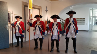 Vier Männer in historischen rot-blauen Militäruniformen, mit Speeren und einer Flagge, stehen in einer hellen Halle.