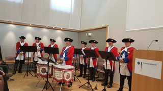 Eine Gruppe Männer in historischen rot-blauen Uniformen mit Dreispitzhüten, Trompeten und Trommeln, bei einem Auftritt.