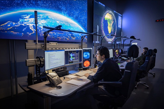 Operatoren überwachen große Bildschirme mit Erd- und Weltraumdaten in einem schwach beleuchteten Kontrollraum.
