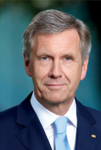 Kopfaufnahme eines lächelnden Mannes mit grauem Haar, blauen Augen, dunklem Anzug und hellblauer Krawatte.