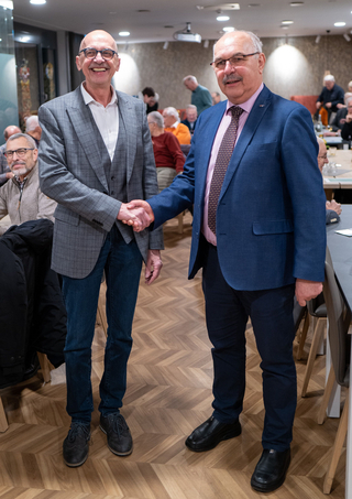 Zwei lächelnde Männer mittleren Alters schütteln sich bei einem Treffen die Hände. Einer trägt einen grauen Anzug, der andere einen blauen.