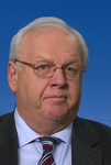 Kopfaufnahme eines älteren weißen Mannes mit kurzen weißen Haaren, Brille, dunklem Anzug und gestreifter Krawatte.