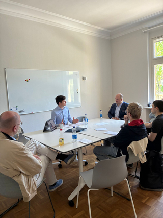 Sechs Männer um einen Konferenztisch in hellem Raum, ein Redner gestikuliert und ein Whiteboard ist im Hintergrund.