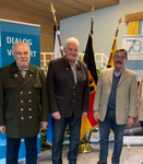 Drei Männer in festlicher Kleidung lächeln in die Kamera, im Hintergrund Flaggen und Veranstaltungsbanner.