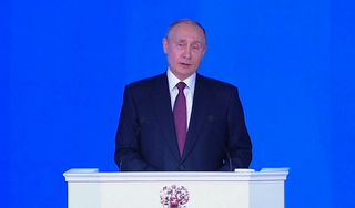 Russischer Präsident Wladimir Putin spricht an einem Rednerpult vor blauem Hintergrund in Anzug und lila Krawatte.