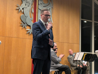 Ein Mann im Anzug spricht in ein Mikrofon auf der Bühne, mit Männern in Militäruniformen und Musikinstrumenten im Hintergrund.