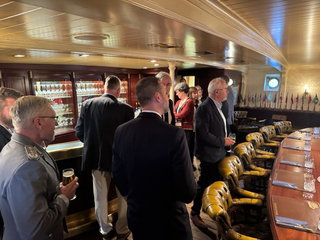 Menschen, darunter ein Mann in Uniform, unterhalten sich im Inneren eines Schiffes mit Bar und langem geschwungenem Tisch.