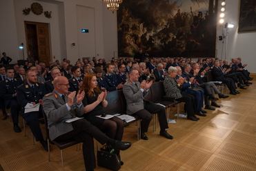 Zuschauer, viele in Uniform, sitzen in einem Saal und klatschen. Im Hintergrund ein großes historisches Gemälde an der Wand.