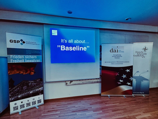 Ein Präsentationsbildschirm zeigt 'It's all about... "Baseline"', umgeben von Bannern von GSP, DAI und der Deutschen Atlantischen Gesellschaft.