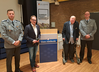 Vier Männer: zwei in Uniform, ein Redner mit Mikrofon, ein älterer Herr mit Rollator, bei einer Reservistenveranstaltung.