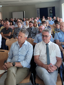 Eine vielfältige Gruppe von Erwachsenen, viele in Uniformen oder Business-Casual, sitzt in einem Hörsaal. Zwei Männer im Vordergrund.