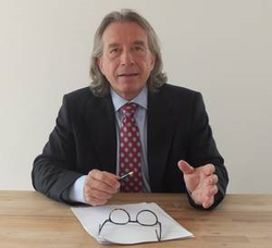Professioneller Mann mit langen Haaren im Anzug am Tisch, hält Stift und gestikuliert erklärend.
