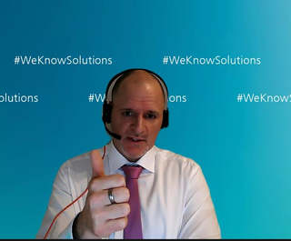 Mann mit Headset und rosa Krawatte zeigt Daumen hoch vor blauem Hintergrund mit '#WeKnowSolutions'.