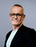 Ein lächelnder Mann mittleren Alters mit kurzen, grauen Haaren und schwarzer Brille, der ein helles T-Shirt und eine dunkle Jacke trägt.