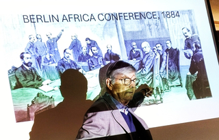 Mann präsentiert eine Folie mit einer Illustration der Berliner Afrika-Konferenz von 1884.