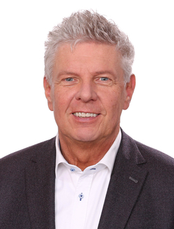 Porträt eines lächelnden Mannes mittleren Alters mit grauem Haar und blauen Augen, in dunklem Blazer und weißem Hemd.