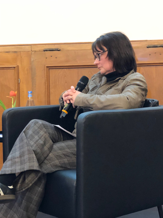 Eine Frau mit Brille und Jacke, die ein Mikrofon hält, sitzt in einem schwarzen Sessel während einer Präsentation.