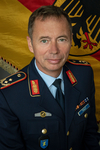 Lachender Brigadegeneral Traut in dunkelblauer deutscher Militäruniform mit Abzeichen und Namensschild, deutsche Flagge im Hintergrund.