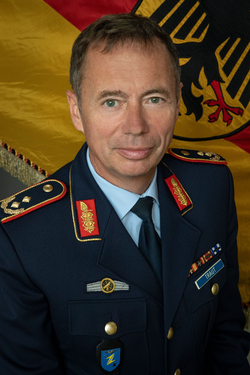 Lachender Brigadegeneral Traut in dunkelblauer deutscher Militäruniform mit Abzeichen und Namensschild, deutsche Flagge im Hintergrund.