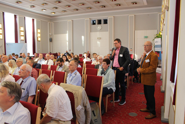 Ein Mann spricht mit einem Mikrofon vor einem sitzenden Publikum in einem Konferenzsaal. Ein weiterer Mann steht daneben.