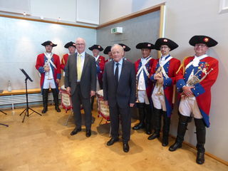 Zwei Männer in Anzügen mit einer Bürgergarde in historischen Uniformen, die Blasinstrumente und Trommeln halten.