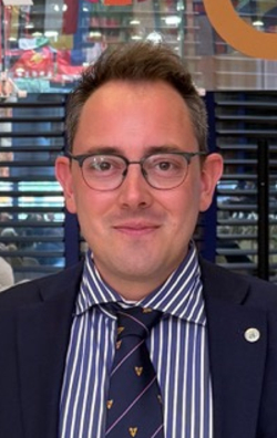 Mann im Anzug und Brille lächelt in einer Lobby mit 'FA 80' und UN SDG Logos auf einer Glaswand, wahrscheinlich bei der FAO.