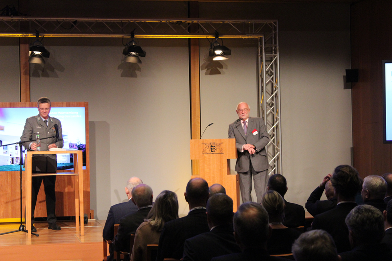 Zwei Männer, einer in Uniform, einer im Anzug, sprechen auf einer Bühne mit Pult und Leinwand vor Publikum bei einer Veranstaltung.