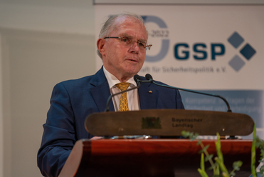 Ein älterer Mann in blauem Anzug und Brille spricht an einem Rednerpult mit Mikrofonen, im Hintergrund ist ein GSP-Logo zu sehen.