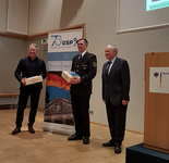 Drei Männer auf Bühne, zwei erhalten Holzschachteln, einer in Uniform, vor GSP-Banner und Rednerpult mit Bundesadler.