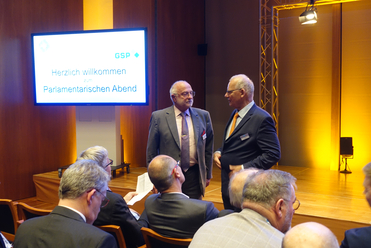 Zwei Männer in Anzügen unterhalten sich vor einer Leinwand mit der Aufschrift 'Herzlich willkommen zum Parlamentarischen Abend' (GSP).