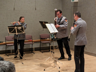 Drei Musiker in militärischen Uniformen spielen Blasinstrumente: eine Frau an der Flöte, ein Mann am Fagott und ein Mann an der Klarinette.