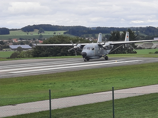 Ein graues Bundeswehr-Transportflugzeug mit Propellern auf einem Rollfeld, dahinter grüne Hügel und ein Ort unter wolkigem Himmel.