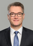 Lächelnder Mann im dunklen Anzug, blauer Krawatte und Brille, vor grauem Hintergrund.