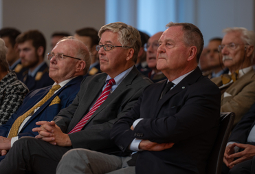Drei Männer in Anzügen und Krawatten, zwei mit Brille, sitzen aufmerksam im Publikum einer Veranstaltung.