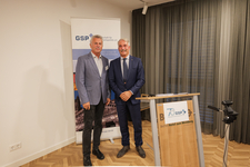 Zwei Männer in Anzügen lächeln vor einem GSP-Banner und Rednerpult bei einer Veranstaltung.