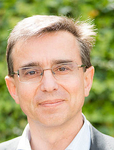 Lächelnder Mann mit Brille und kurzen, strähnigen Haaren im Porträt vor unscharfem grünem Hintergrund.