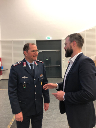 Zwei Männer unterhalten sich drinnen; einer in dunkler Uniform mit Abzeichen und Headset, der andere im Anzug mit Bart.