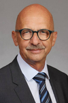 Professionelles Porträt eines lächelnden glatzköpfigen Mannes mit dunkler Brille und Schnurrbart, im Anzug mit gestreifter Krawatte.