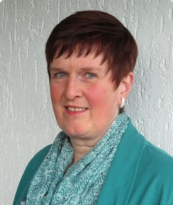 Porträt einer lächelnden Frau mit kurzen rotbraunen Haaren, blauen Augen, türkisfarbenem Oberteil und gemustertem Schal.