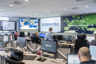 Frontex-Lagezentrum mit Personal an Schreibtischen und großen Videowänden, die Karten und das Agenturlogo anzeigen.