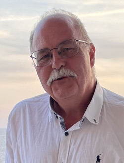 Porträt eines älteren Mannes mit Brille und Schnurrbart in weißem Poloshirt, vor sanftem Himmel und Meer.