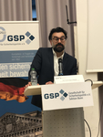 Ein Mann, Ali Fathollah-Nejad, spricht bei einem GSP-Event an einem Rednerpult vor deutscher Flagge und Reichstags-Abbildung.