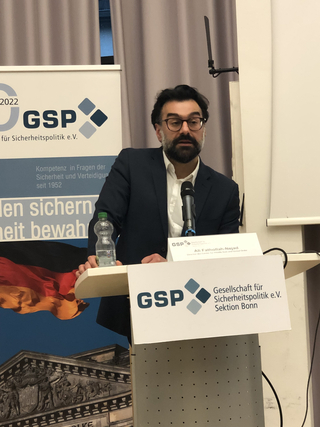 Ein Mann, Ali Fathollah-Nejad, spricht bei einem GSP-Event an einem Rednerpult vor deutscher Flagge und Reichstags-Abbildung.
