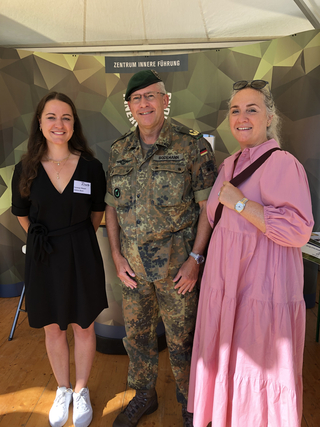 Drei lächelnde Personen, zwei Frauen und ein Mann in Uniform, stehen vor einem Banner des 'ZENTRUM INNERE FÜHRUNG'.