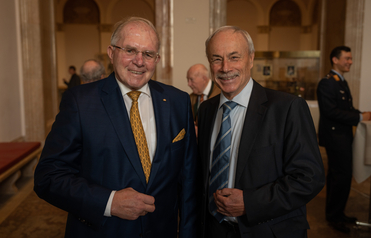 Zwei lächelnde ältere Herren in Anzügen, einer mit goldener, der andere mit blauer Krawatte, posieren bei einer Veranstaltung.