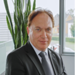 Professionelles Porträt eines lächelnden Geschäftsmannes in dunklem Anzug mit Pflanze im Hintergrund.